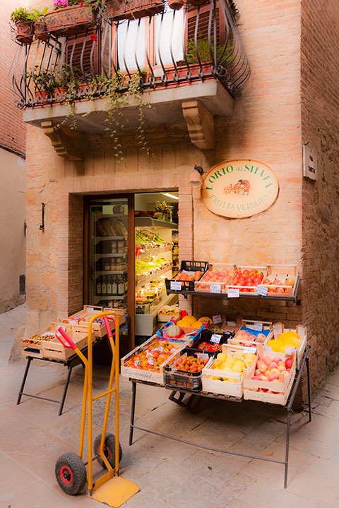 bright yellow hand truck sits outside a tiny Tuscan neighborhood market in Pienza, Italy as the vegetable deliveryman finalizes the transaction inside