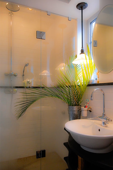 palm leaves in corrugated steel vase set against shower glowing softly through its minimal glass wall
